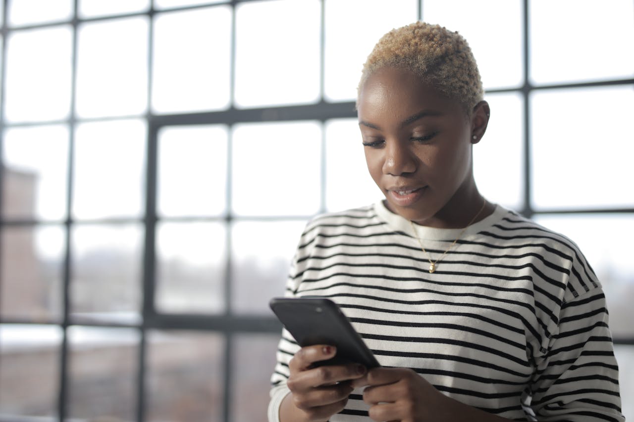 Home Young African American woman texting on smartphone by a window with natural light.